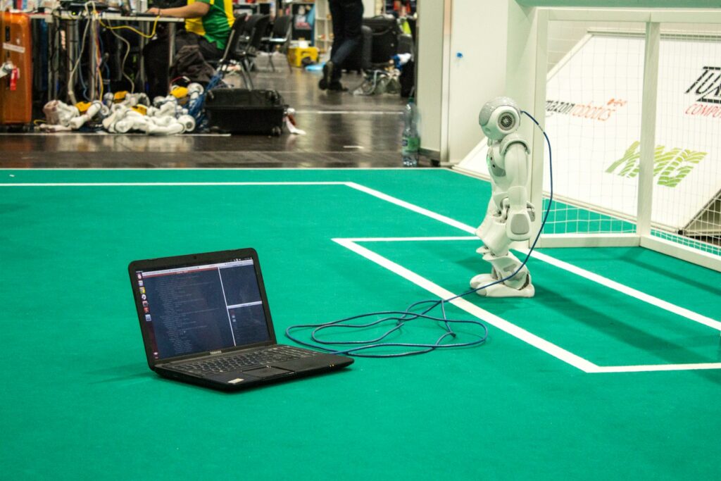 Robot on soccer field connected to laptop with coding visible. Innovative technology.